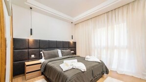 A modern bedroom with a dark headboard, a wooden nightstand, a fluffy white rug, and a large window with sheer curtains.
