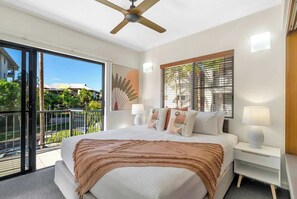 A bedroom with a bed, bedside tables, a ceiling fan, a window with blinds, and a balcony with a view of greenery and buildings.