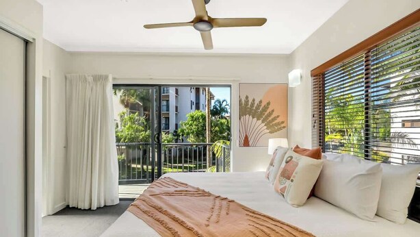 A hotel room with a bed, a ceiling fan, a balcony with a view of buildings and greenery, and a window with blinds.