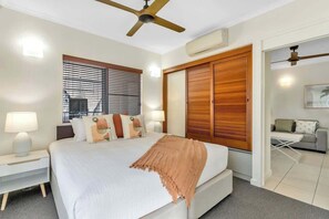 A hotel room with a bed, bedside lamps, a ceiling fan, a window with blinds, a wooden wardrobe, and a small seating area with a table and a sofa.