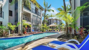 A pool area with lounge chairs, palm trees, and a building with balconies.