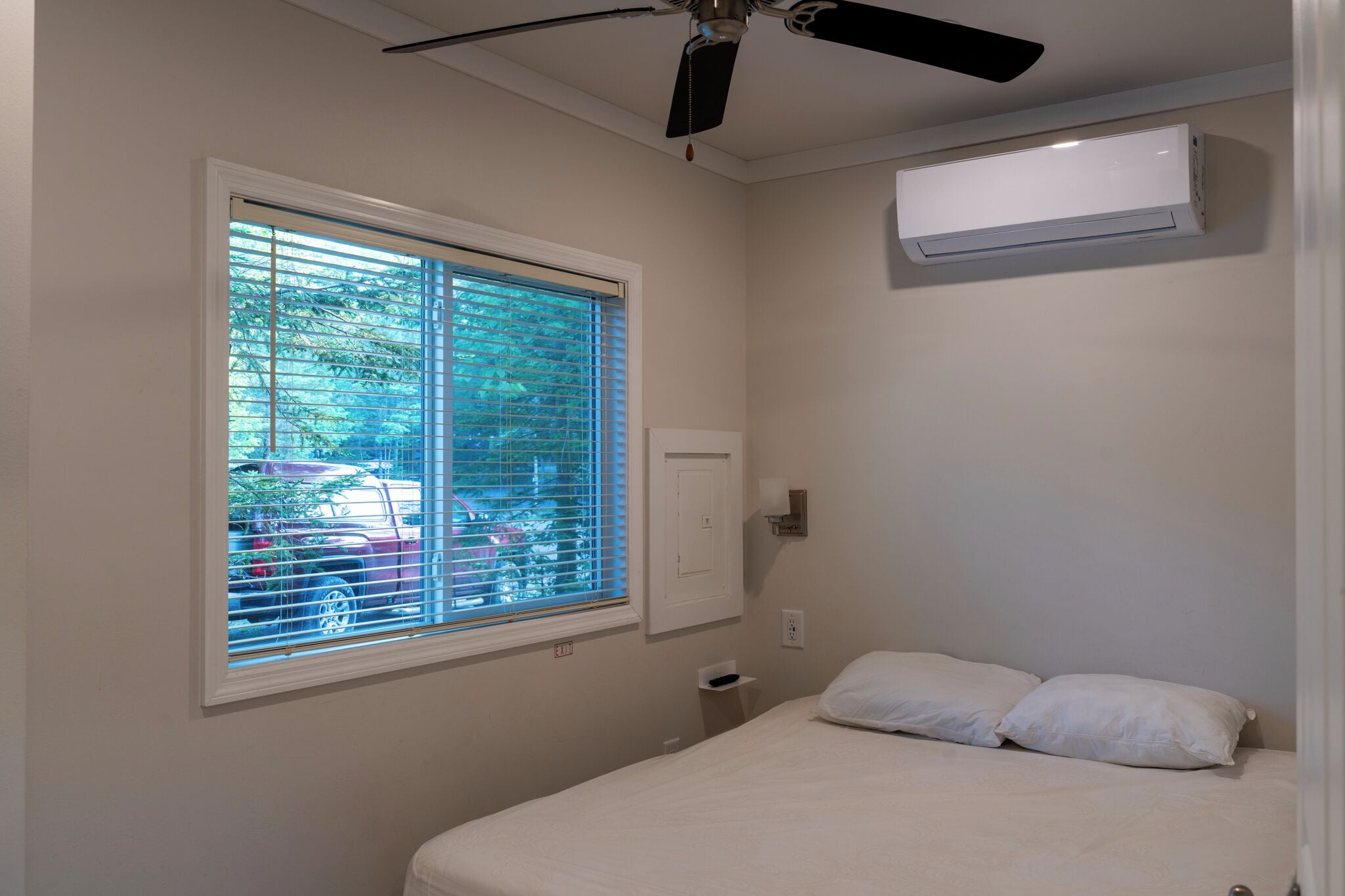 A bedroom with a bed, a ceiling fan, an air conditioning unit, a window with blinds, and a view of the outside.