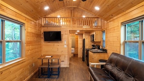 A cabin interior with a loft bedroom, a dining area, a kitchen, and a living room with a leather couch. - HTR Adirondacks (Thendara)