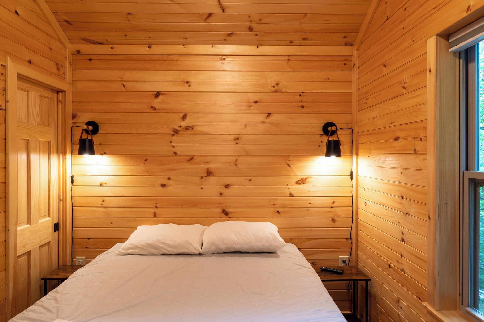 Una habitación con paneles de madera, una cama, dos lámparas de pared y un ventanal.