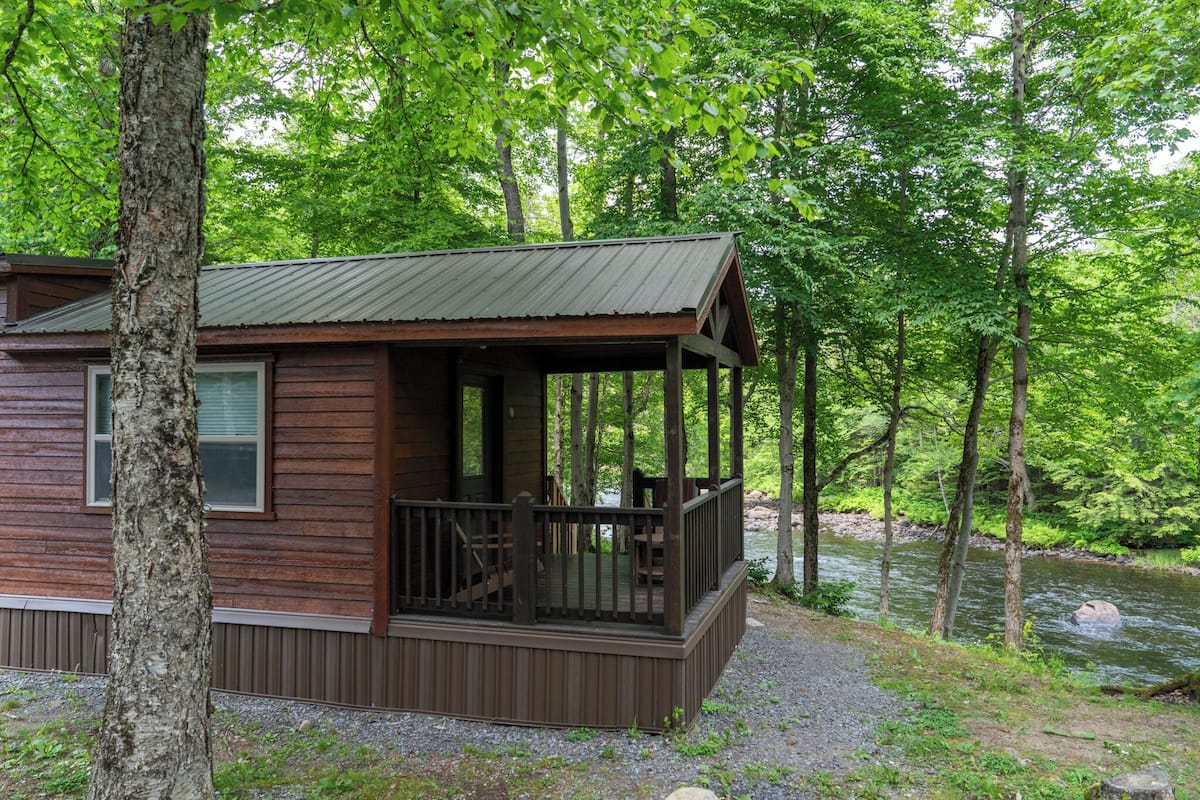 A wooden cabin with a porch, surrounded by a forest and a stream.
