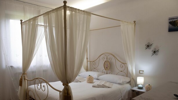 A four-poster canopy bed with a white bedspread and pillows, a bedside table with a lamp, and a wall-mounted picture. - Double Room with Pool View (Lazio)