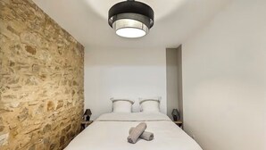 A bedroom with a stone wall, a bed with white linens, two pillows, a bedside table with a lamp, and a modern ceiling light fixture.