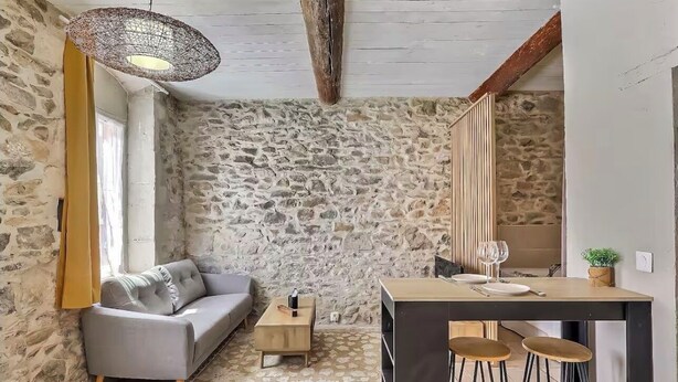 A cozy room with a stone wall, a grey sofa, a wooden coffee table, a hanging light fixture, and a small bar area with stools.
