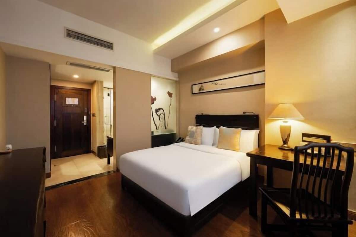 A hotel room with a bed, a desk, a chair, a lamp, and a painting on the wall.