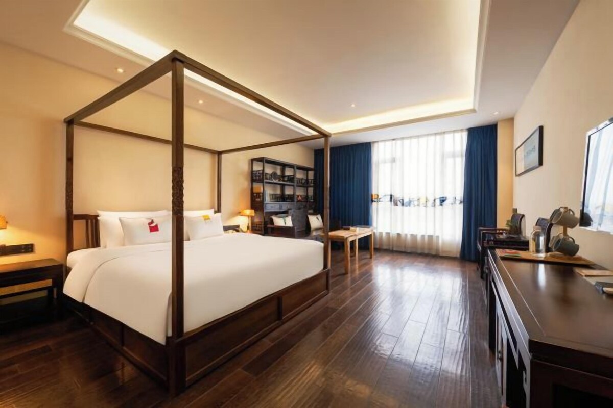 A four-poster bed with a wooden frame, a bedside table with a lamp, a bookshelf, and a large window with curtains.