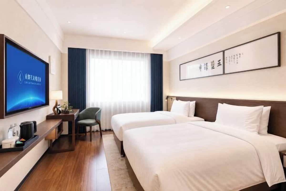 A hotel room with two beds, a TV, a desk, and a window with curtains.