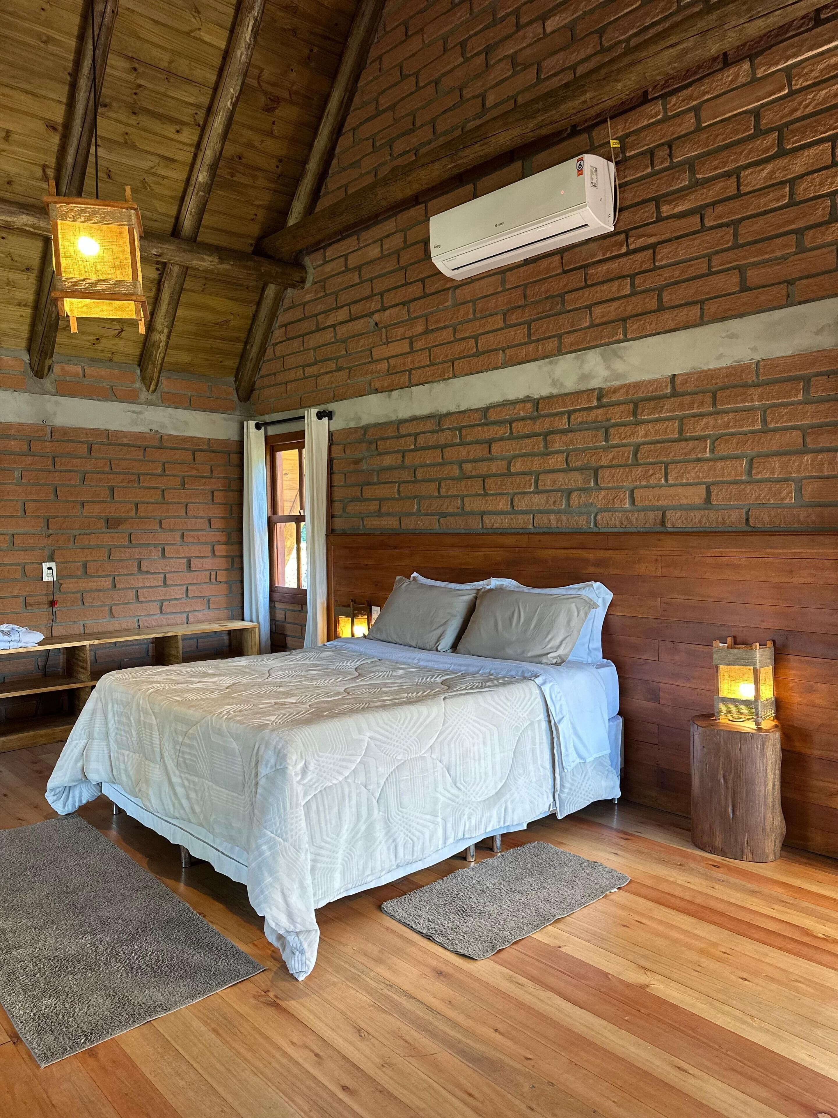 A bedroom with a wooden ceiling, brick walls, a bed with a white bedspread, a nightstand, a lamp, and an air conditioning unit.