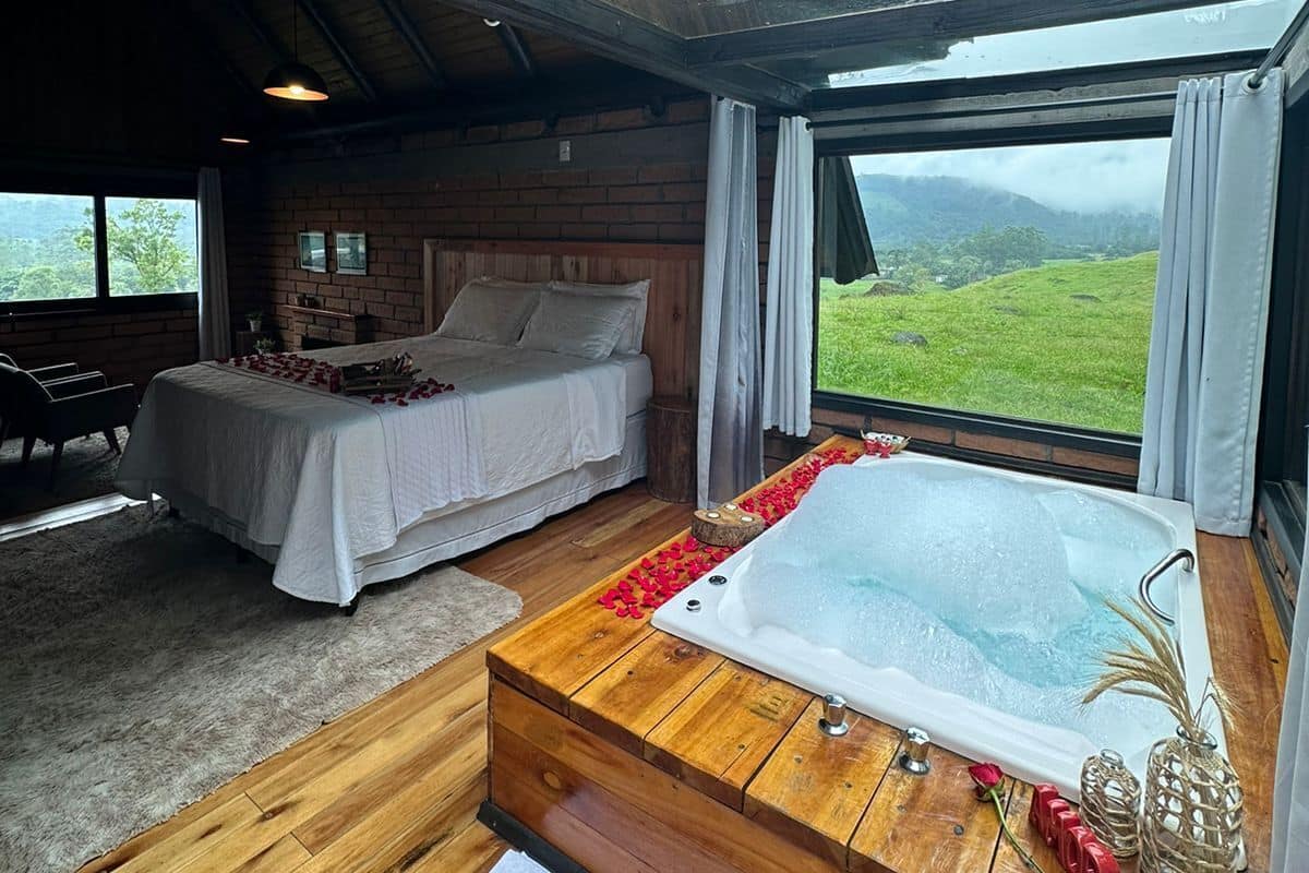 A bedroom with a large window, a wooden floor, a bed, a sofa, and a bathtub.