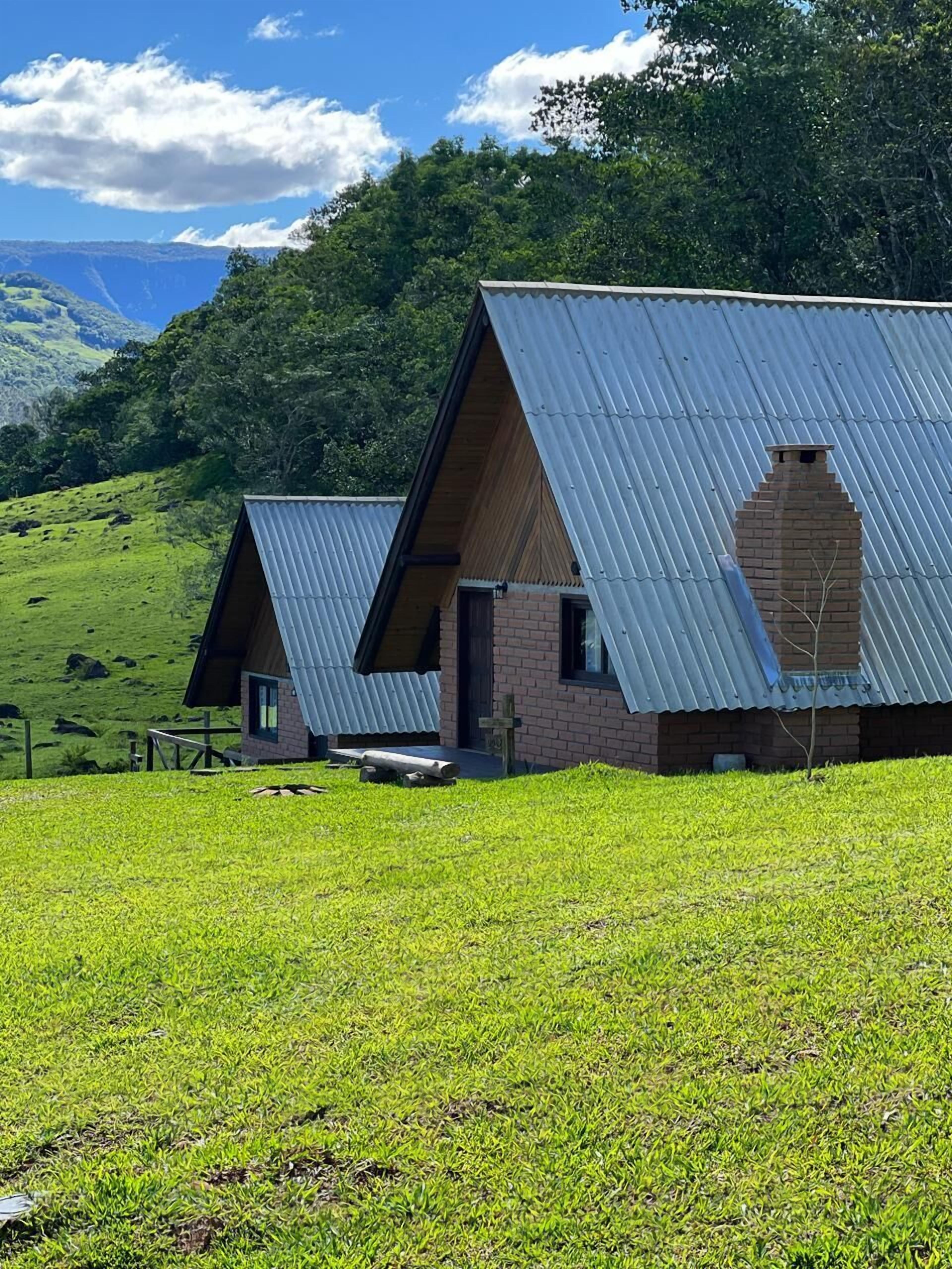 A rustic cabin with a sloped metal roof, surrounded by lush green fields and a mountainous backdrop.
