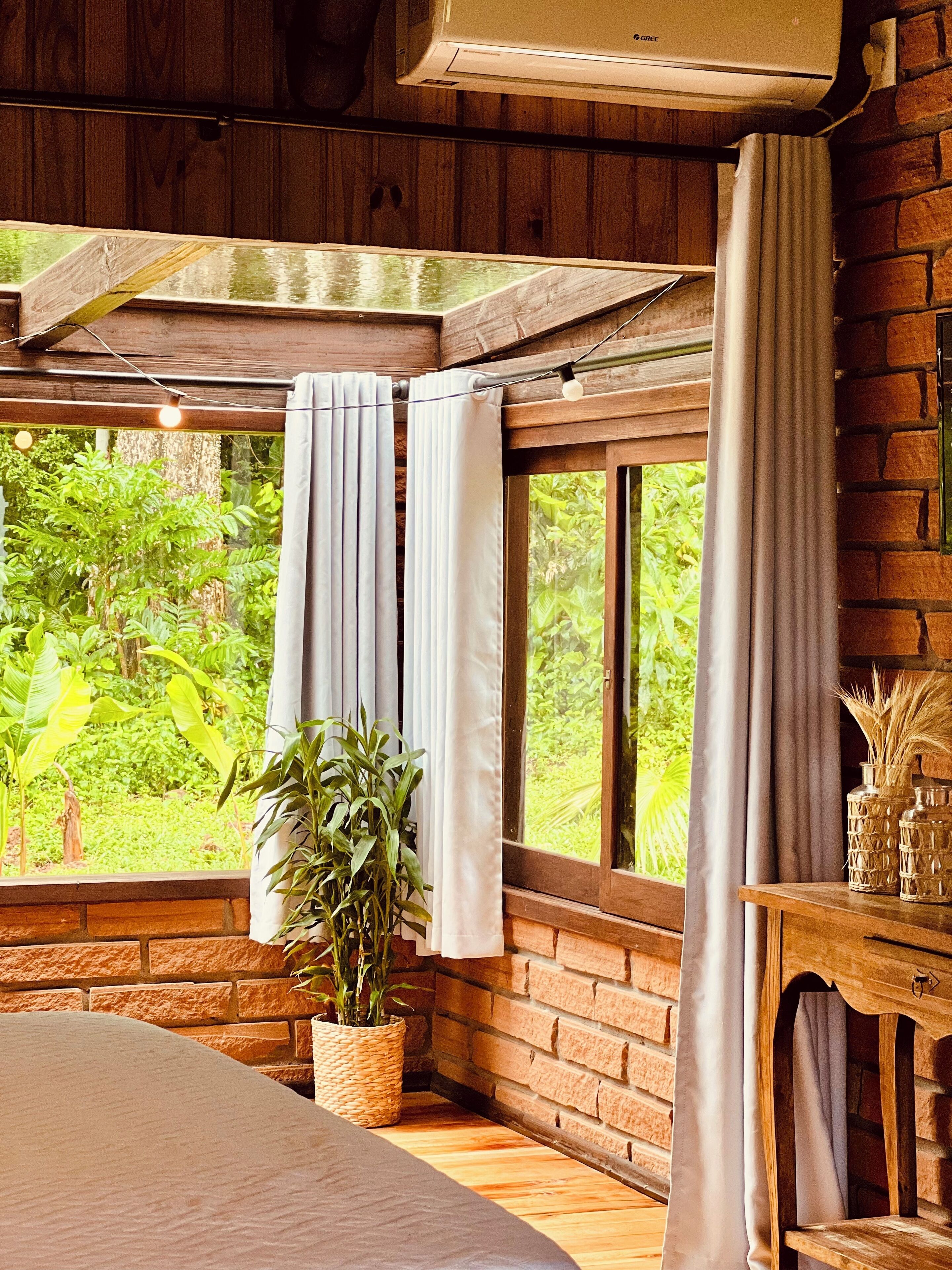Una habitación de madera con una cama, una planta en maceta, una ventana con cortinas y una pared de ladrillo.