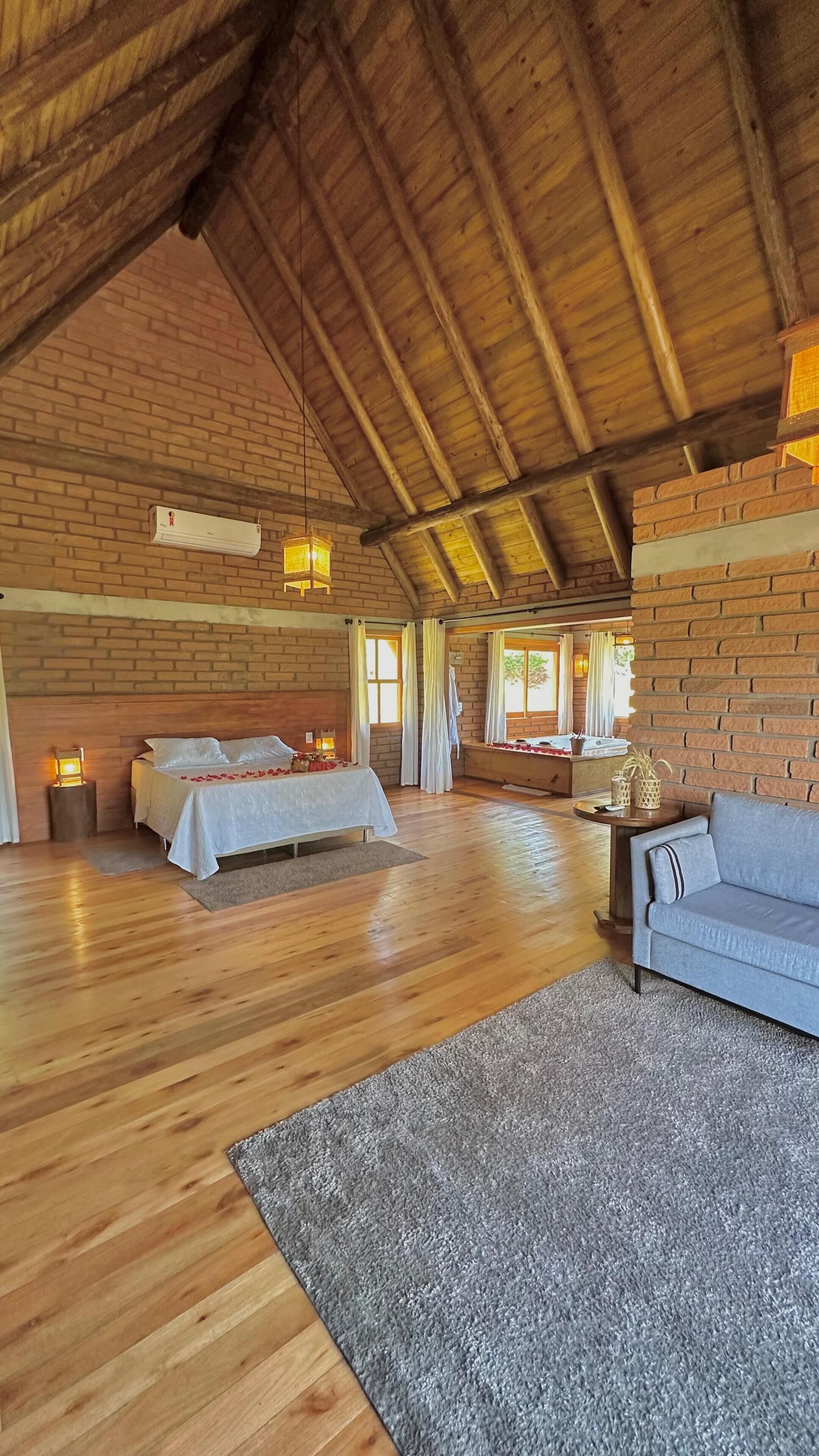 Una habitación amplia con un techo alto de madera, una cama con colcha blanca, un sofá gris y una mesita de noche con una lámpara.