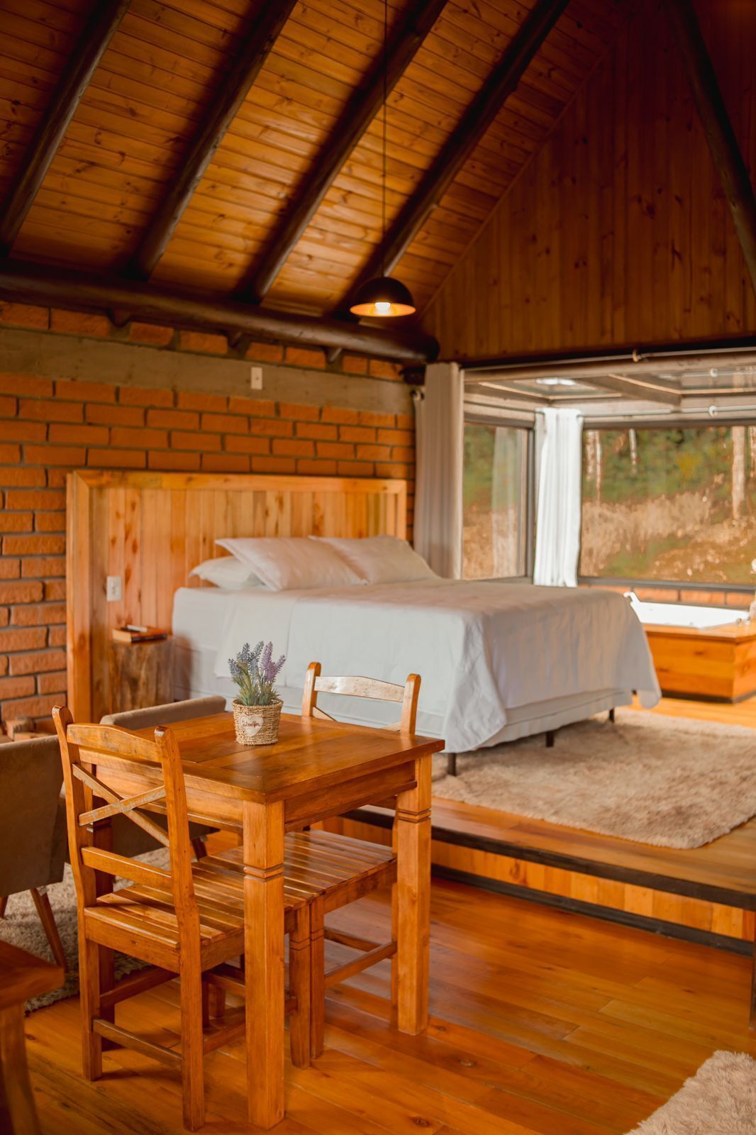 A wooden cabin bedroom with a bed, a dining table with chairs, and a window overlooking a natural landscape.