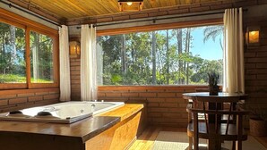 A wooden cabin interior with a hot tub, a dining table, and a view of the forest. - Maria Rita dos Canyons (Praia Grande)