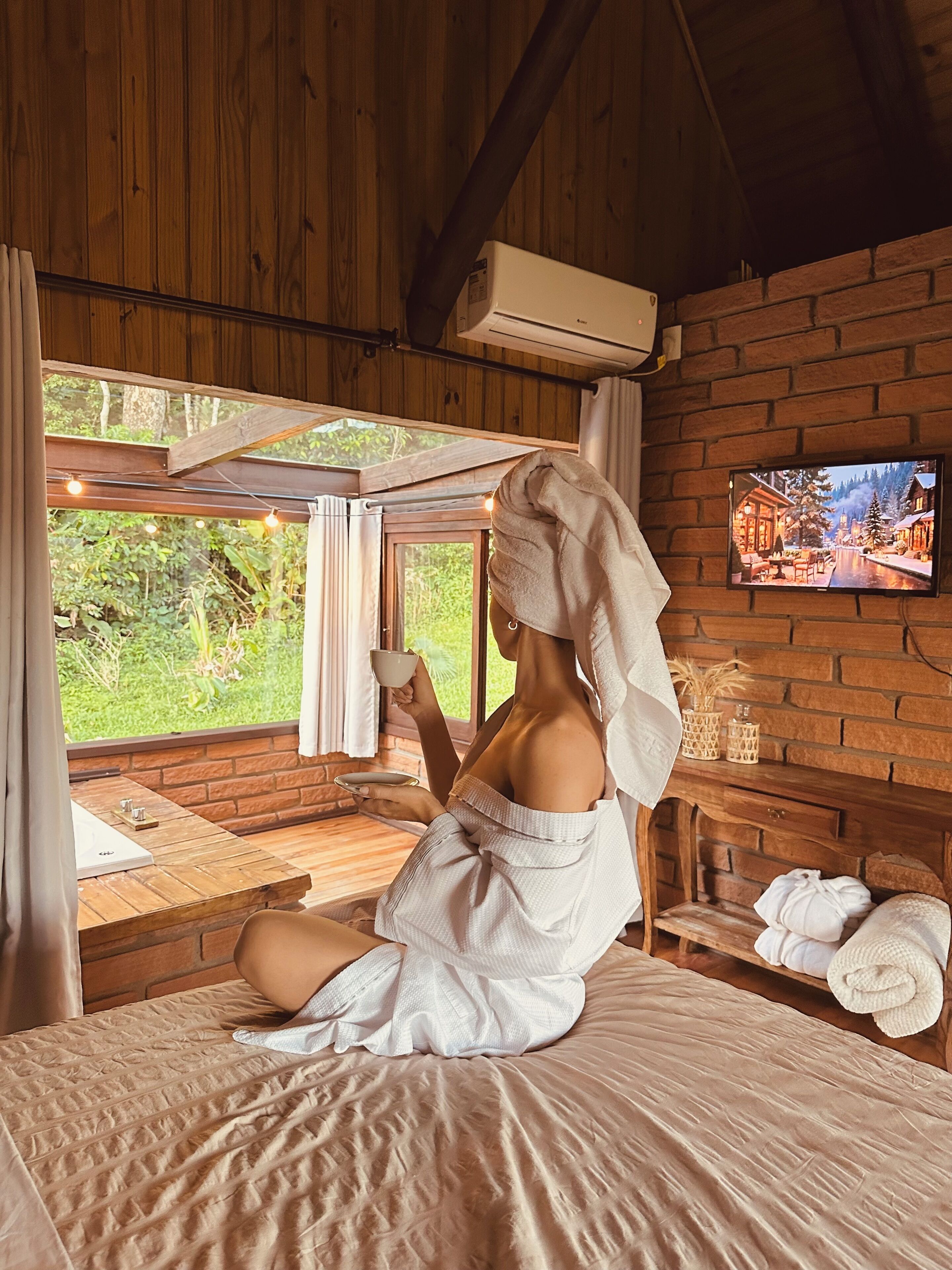 A room with a wooden ceiling, a brick wall, a TV mounted on the wall, a window with curtains, and a bed with a towel on it.