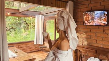 A room with a wooden ceiling, a brick wall, a TV mounted on the wall, a window with curtains, and a bed with a towel on it.