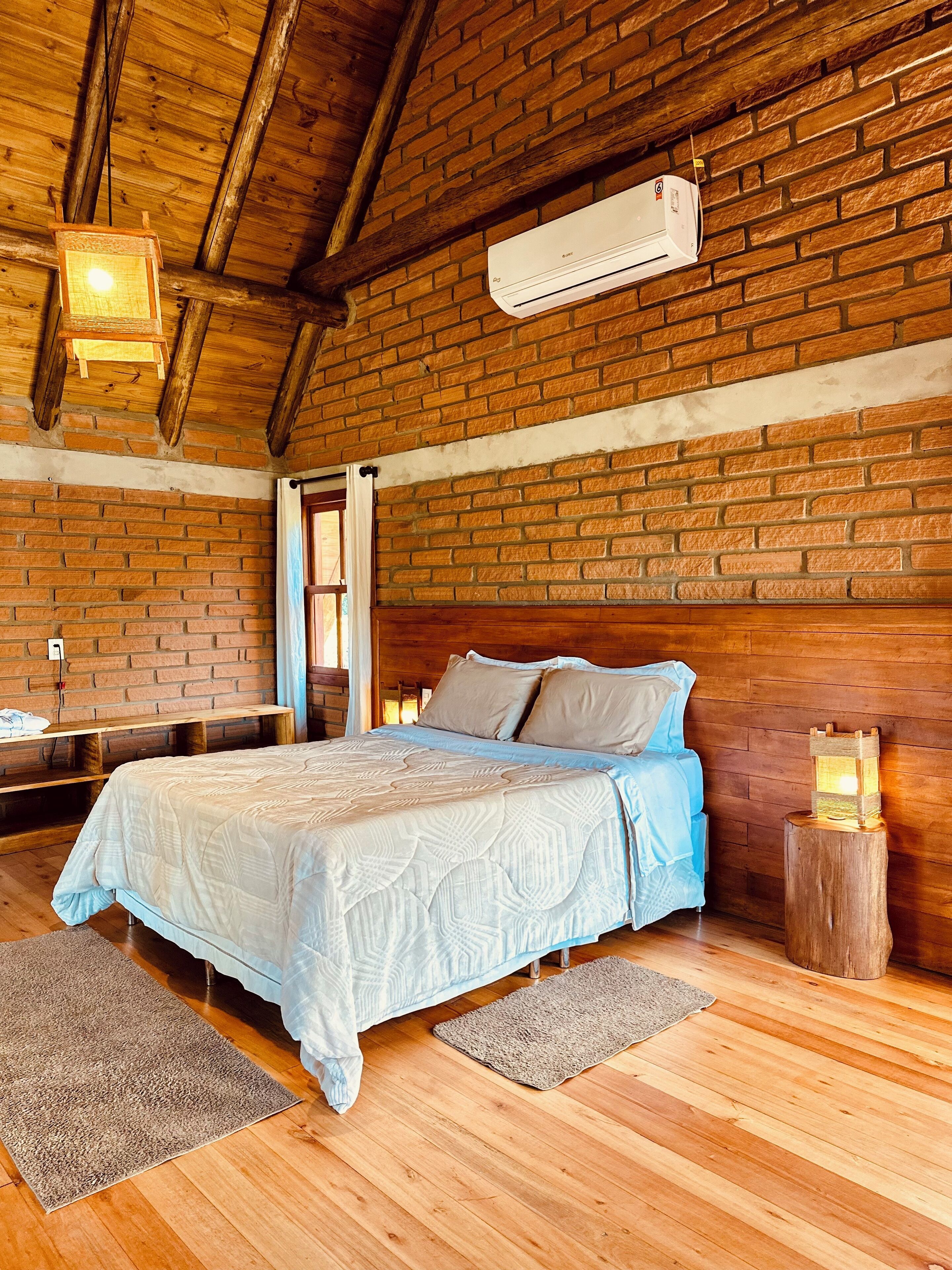 Une chambre avec un plafond en bois, des murs en briques, un lit recouvert d’une courtepointe bleue et blanche, une table de chevet et une petite table de nuit avec une lampe.