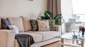 A living room with a beige sofa, patterned cushions, a glass coffee table, and large green plants. - Sunflower Stay by Homie (Santa Cruz)