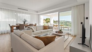 A modern living room with a sofa set, a coffee table, and a TV. There is a dining area with a table and chairs. The room has large windows with curtains, offering a view of the outdoors.