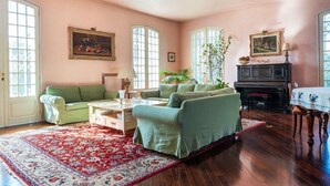 A cozy living room with a green sofa, a coffee table, and a fireplace. There are two windows with curtains, a door, and a red patterned rug on the floor.