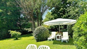 A garden patio with white chairs and a table, surrounded by lush greenery and trees.