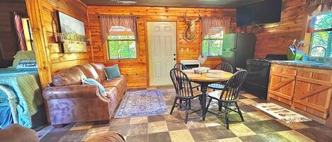 A cozy cabin interior with a brown leather couch, a dining table with chairs, a kitchen area, and a checkered floor.