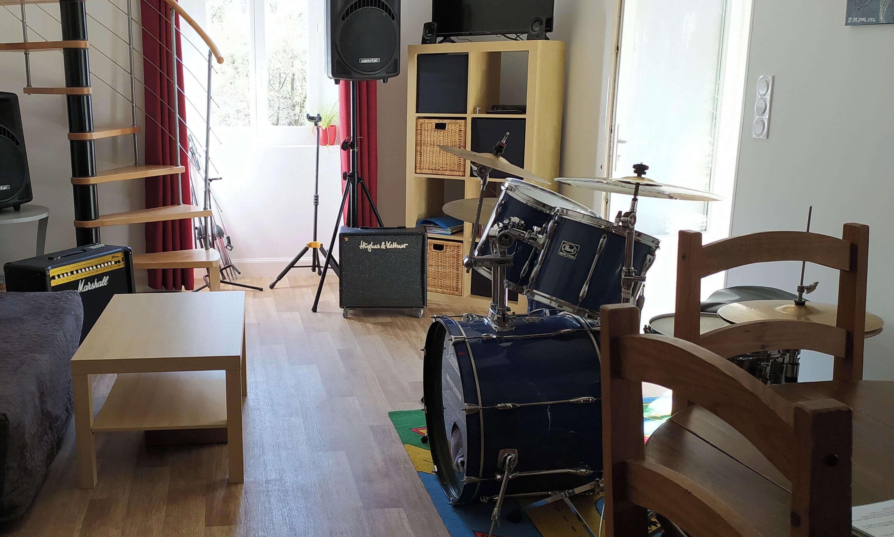 A room with a drum set, a Marshall amplifier, a bookshelf, and a window with curtains.