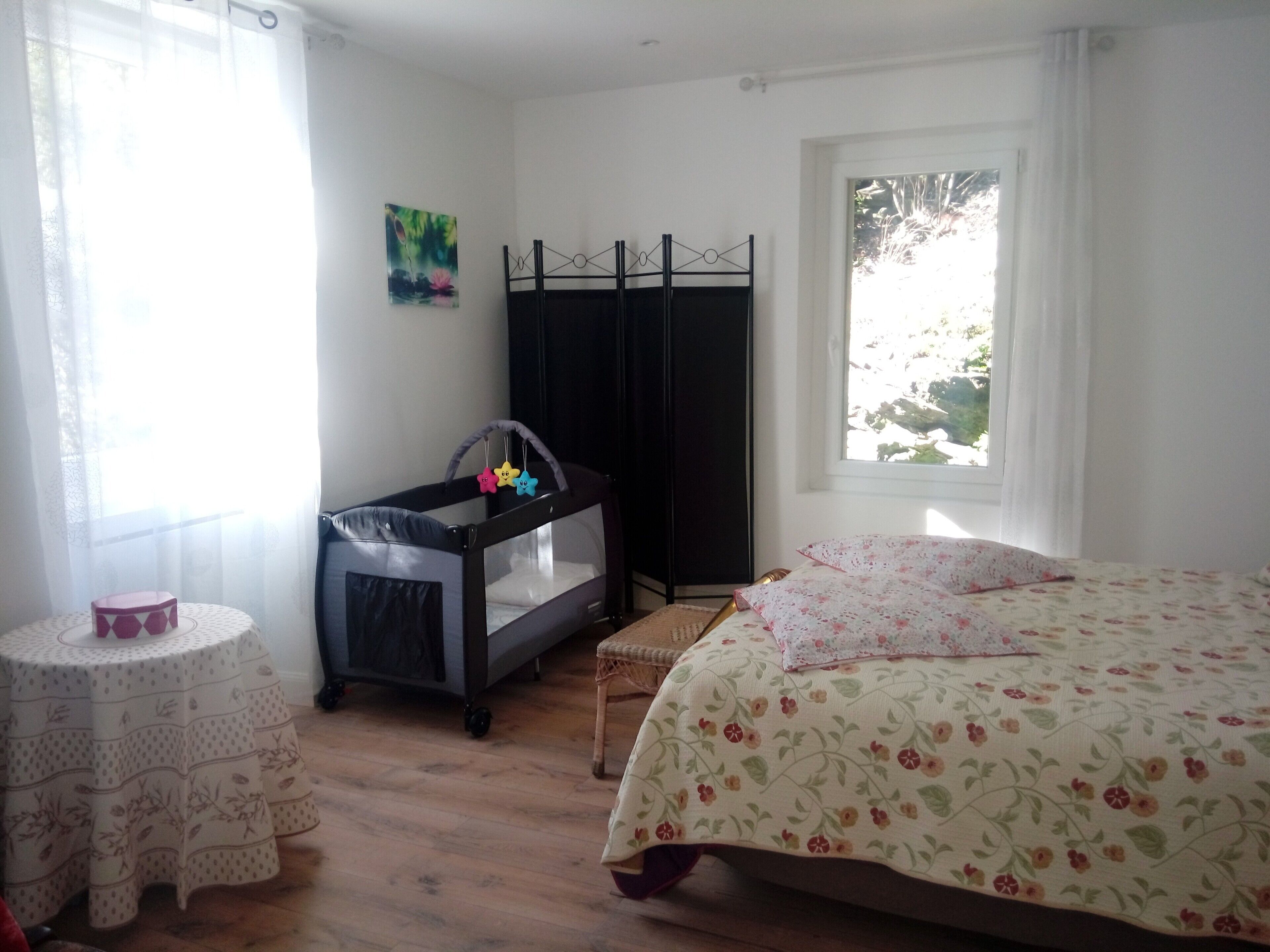 Un dormitorio con una cuna, una mesita redonda pequeña, una cama con ropa de cama floral, una ventana con vistas y un cuadro en la pared.