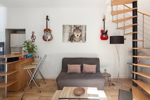 A modern living room with a grey sofa, wooden shelves, a dining area with a table and chairs, and a spiral staircase.
