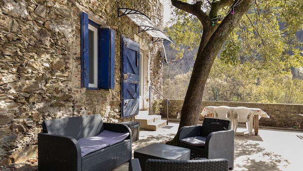 Un área de estar al aire libre con mobiliario de mimbre, paredes de piedra y un árbol que da sombra.