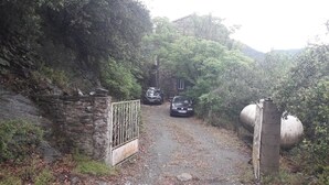Un sendero de grava que conduce a un muro de piedra con una puerta de metal, rodeado de follaje denso y una colina al fondo.