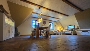 A spacious loft with a high ceiling, wooden beams, a dining area with a table and chairs, a sofa, and a large window providing natural light. - Vacation apartment "Hahnskamp" / 2-6 pers. - Vacation on the farm - (Hamminkeln)