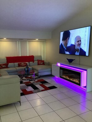 Ein modernes Wohnzimmer mit einem flachbildfernseher an der Wand, einem weißen Sofa mit roten Kissen, einem Glastisch und einem gemusterten Teppich.