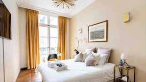 Habitación de hotel con una cama grande, una mesita de noche con lámpara, un cuadro en la pared y vistas a la ciudad desde la ventana.