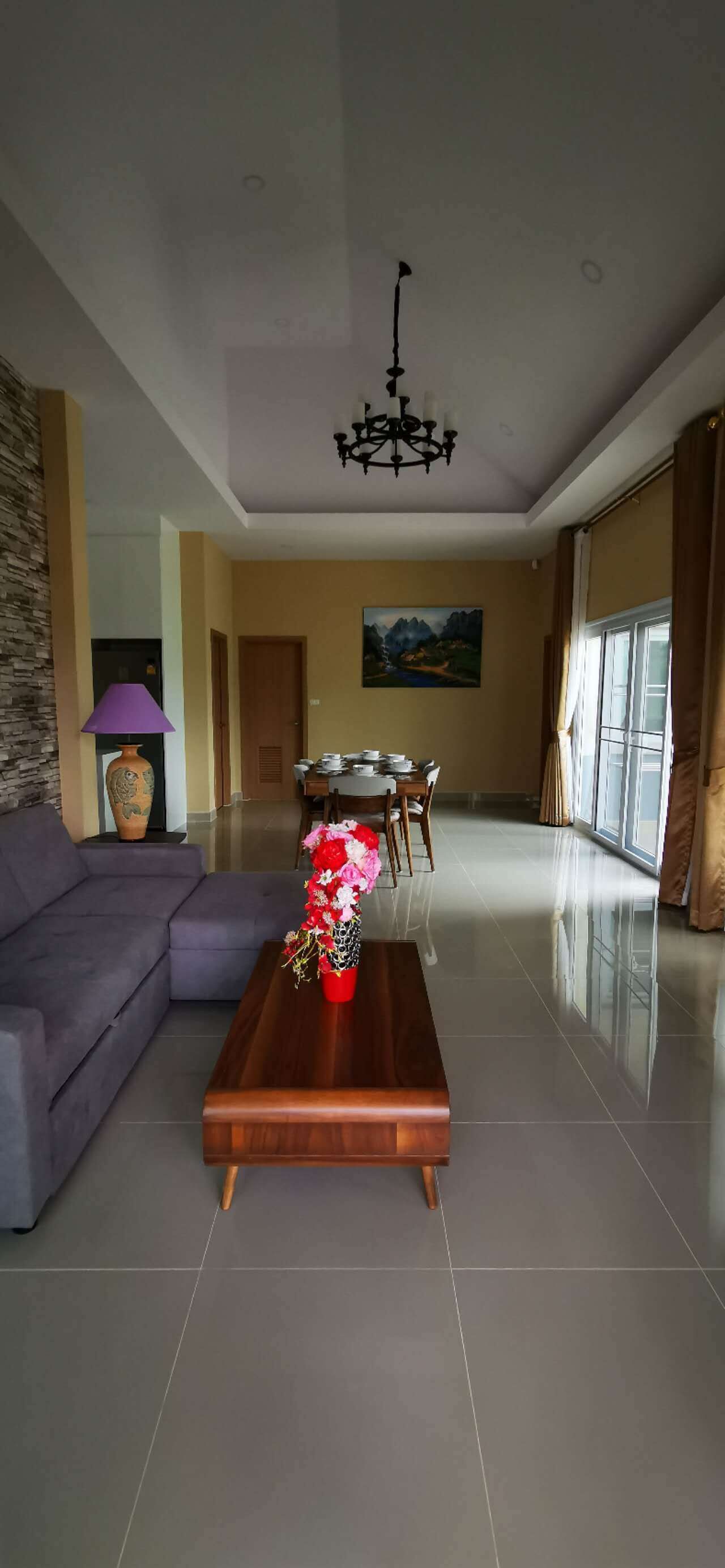 A spacious living room with a modern sofa, a wooden coffee table with a floral centerpiece, a dining area with a table and chairs, and a large window providing natural light.