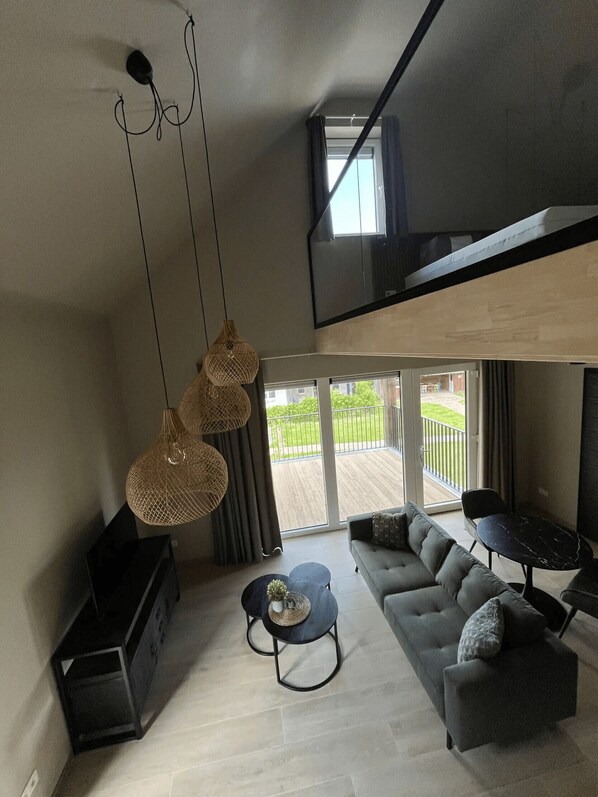 Ein modernes Wohnzimmer mit einer Couch, einem Couchtisch und Pendelleuchten. Es gibt einen Balkon mit Blick auf Grünflächen.