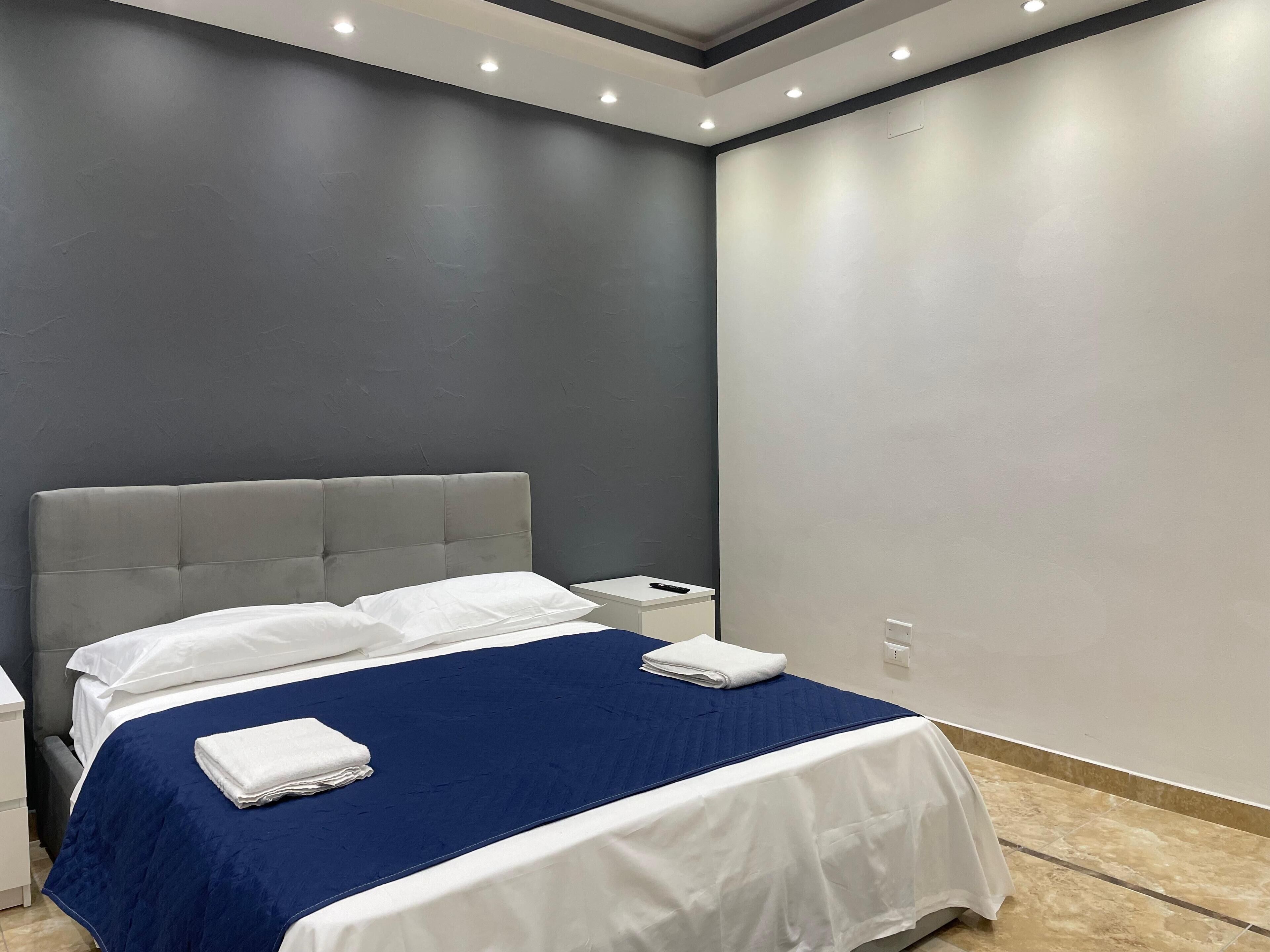 Un dormitori modern amb un capçal de llit tapissat de gris, un edredó blau, almoines blanques i una taula de nit blanca amb un comandament a distància.