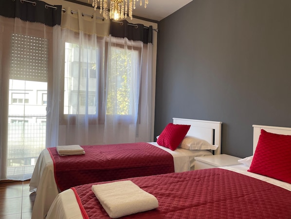 Habitación de hotel con dos camas, un ventanal con cortinas translúcidas, un candelabro y vistas al exterior.