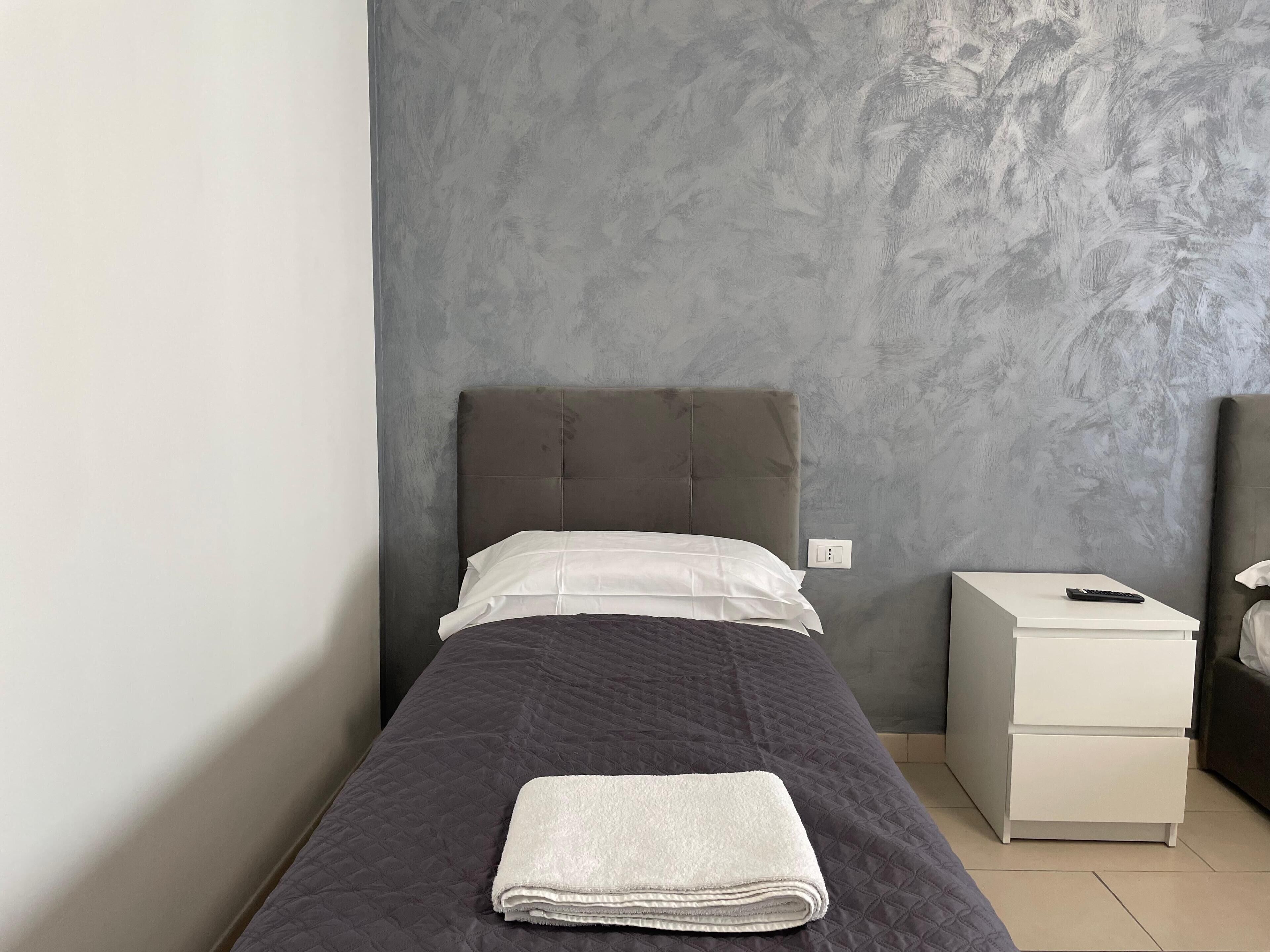 A single bed with a grey headboard, a white pillow, and a folded white towel on the bed. There is a white bedside table with a remote control on it. The wall behind the bed has a textured grey finish.