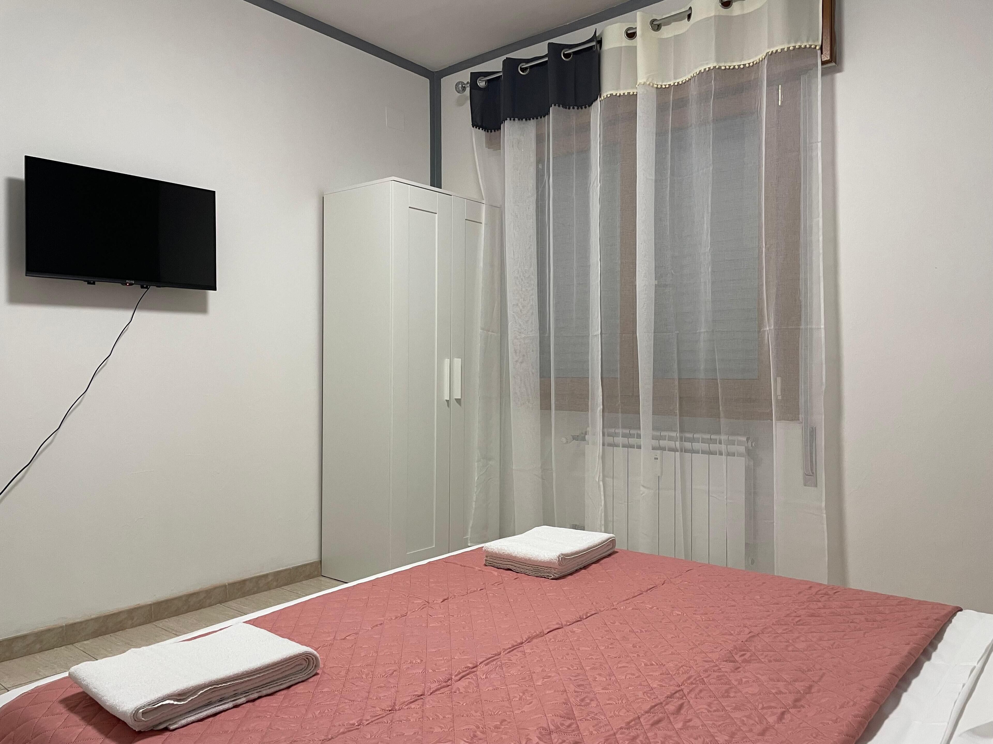 Un dormitorio con cama, televisor montado en la pared, armario y ventana con cortinas ligeras.