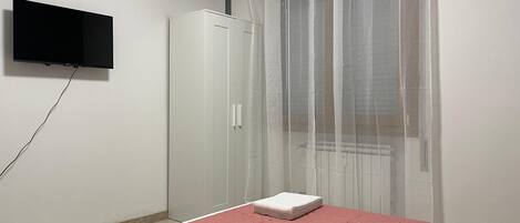 Un dormitorio con cama, televisor montado en la pared, armario y ventana con cortinas ligeras.