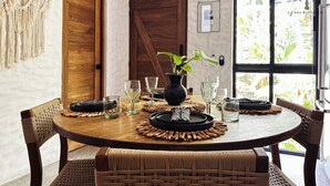 Une table de salle à manger ronde en bois, avec des chaises en osier, une plante en pot et un plateau noir contenant des couverts.