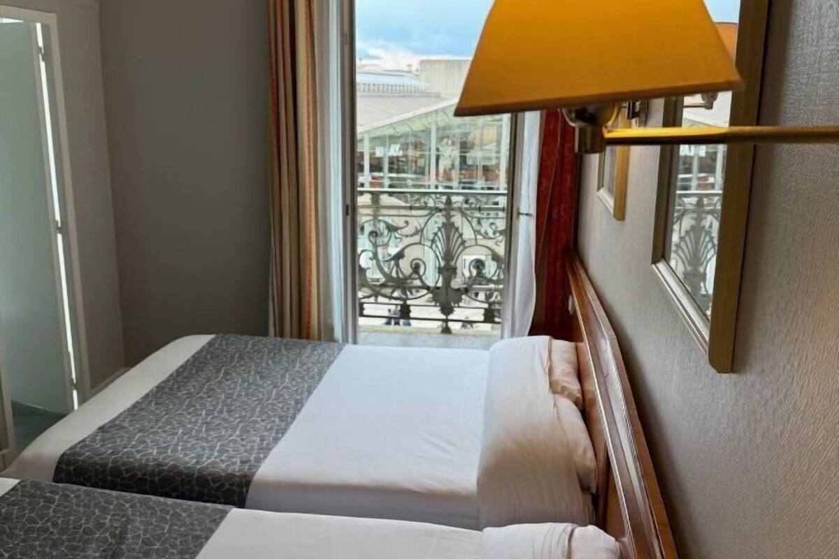Una habitación de hotel con dos camas, un ventanal con vistas a un balcón y una lámpara en la pared.