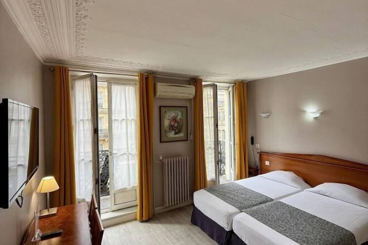 Habitación de hotel con dos camas, un aparador de madera, un escritorio con lámpara y un ventanal con cortinas.