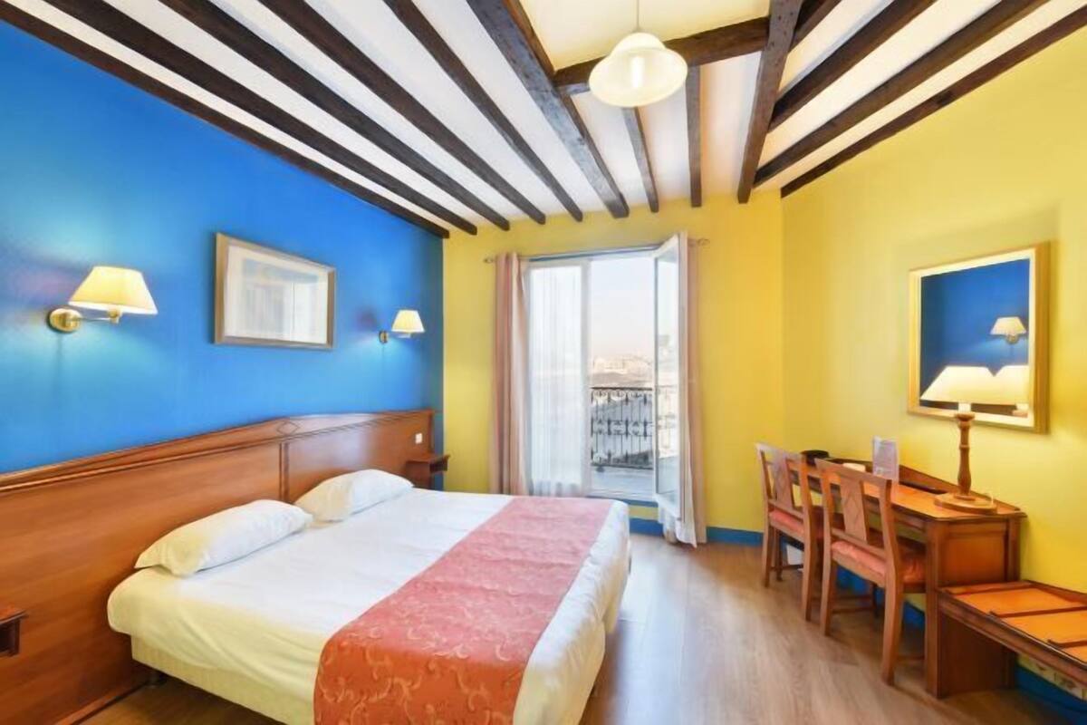 Habitación de hotel con cama, escritorio y silla. Se ve una ventana con vistas al exterior.