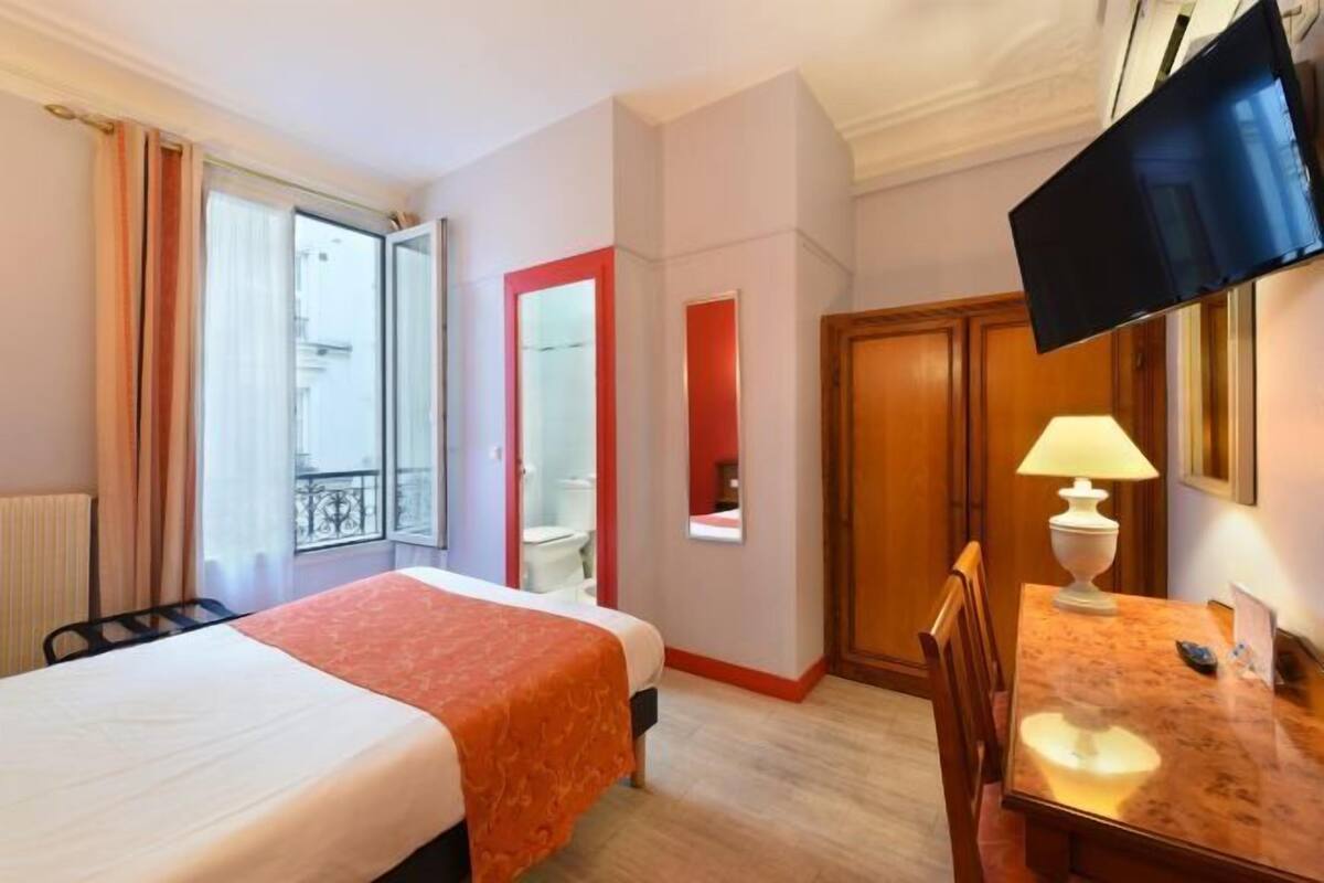 Habitación de hotel con cama, escritorio, silla, televisor y ventana con cortinas.